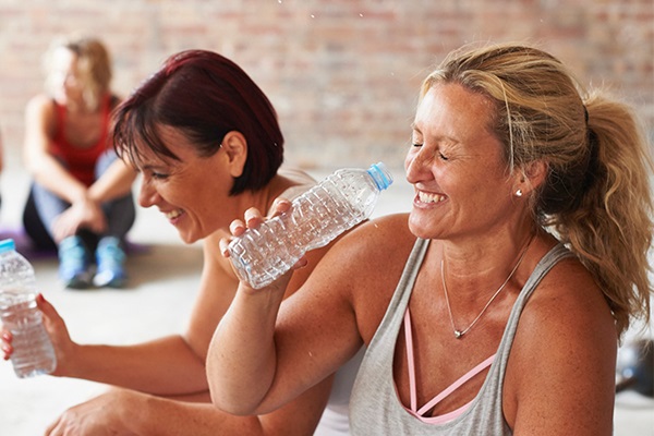 Two friends sitting on the floor at a gym, holding water bottles and laughing.