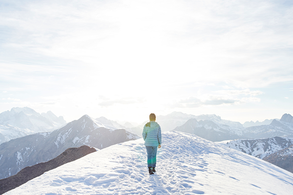 A woman in a green jacket walking along the top of a snowy mountain, looking out at a sunny sky and surrounding mountains.