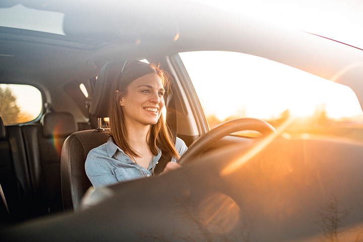 A woman driving a car and smiling, with sun shining through the windows of the car.