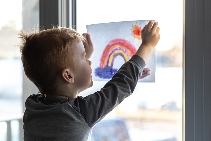 Young boy inside his home, taping a drawing of a rainbow to his window.