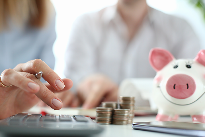 A couple sitting at a table, on which there is a calculator, stacks of coins, and a smiling piggy bank. The woman is punching the calculator buttons while holding a pen.