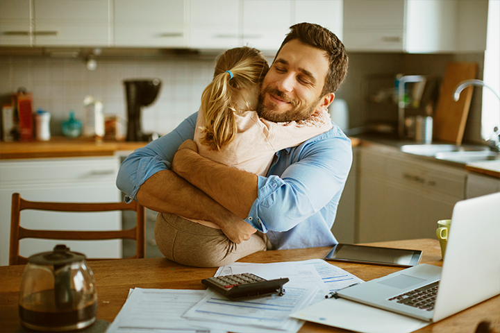 Man sitting at kitchen table with computer and work-related documents, hugging his daughter who is sitting on his lap.