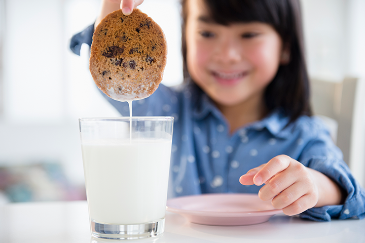 A young girl dipping a chocolate chip cookie in a glass of milk and smiling.