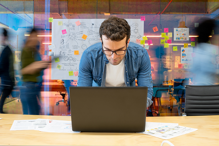 A man standing in front of a laptop computer on a table, with a buzz of activity happening behind him in an office.