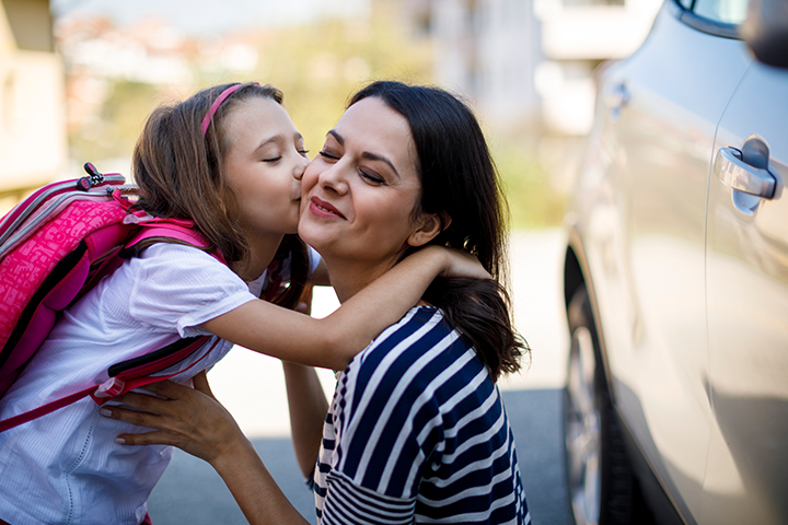 A mom, crouched down in front of her car, and her daughter wearing a pink backpack leaning in for a hug and kiss on the cheek.