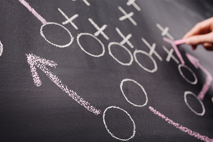 A chalkboard scattered with X's and O's representing team players, and a woman's hand drawing arrows around the board depicting play moves.