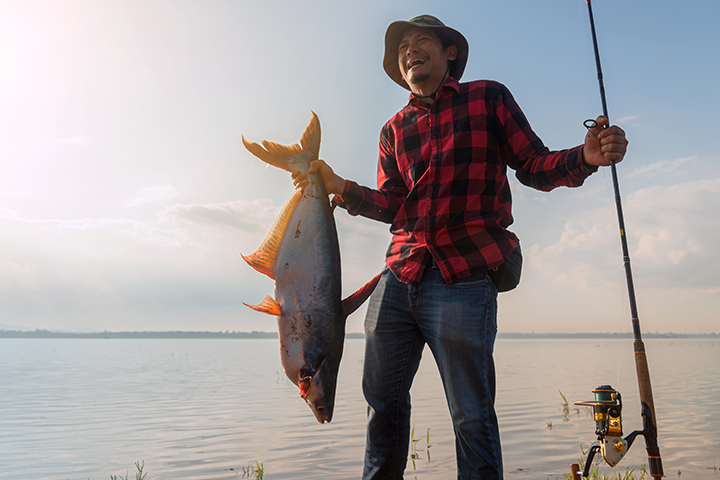 A man with a huge smile, wearing a plaid shirt and fisherman's hat, holding a fishing pole and a huge fish, standing in front of a lake.