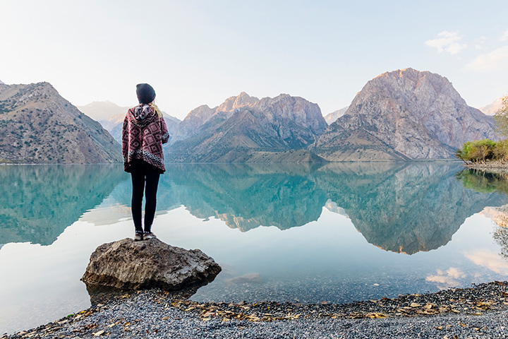 A woman dressed in warm clothing looking across a crystal-clear lake showing reflections of the surrounding mountains.