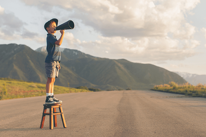 A young boy standing on a stool on a long dirt road, shouting through a mega phone.