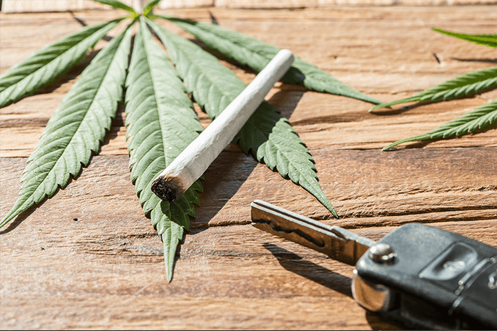 A cannabis leaf lying on a wooden table, with a rolled joint over top, and a set of car keys next to it.