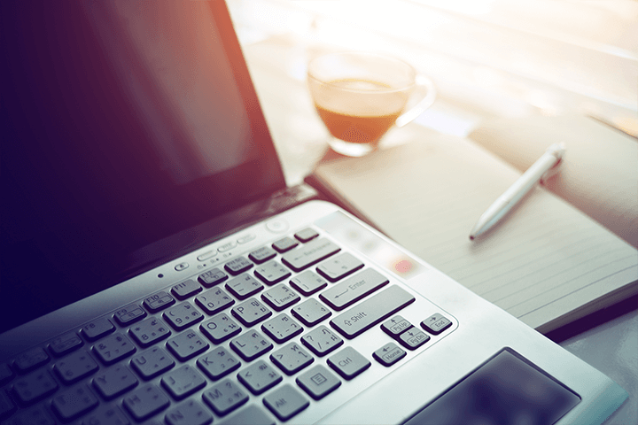 A laptop computer open, with a notebook and pen next to it, and a cup of coffee.