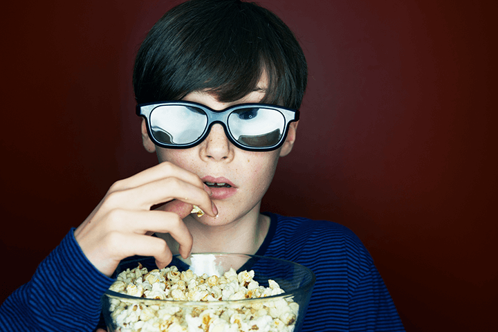 A young boy wearing 3D glasses, eating popcorn with a look of suspense and anticipation