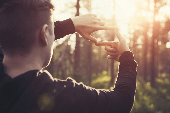 A man looking at a forest through his hands which are shaping a square, with sun shining in background.