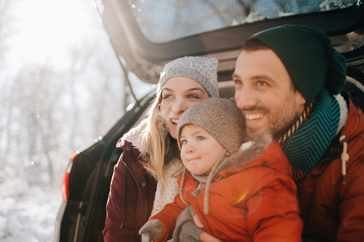 A young couple sitting in the open trunk of their minivan, holding a small child on their lap. They are all bundled up in winter wear, and the background is a wintery forest with the sun shining through.