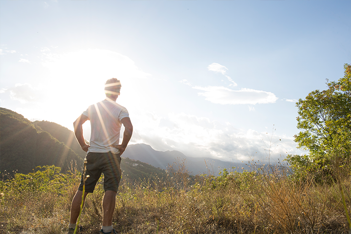 A young man standing on top of a grassy hill, looking out at a great expanse, with sun beams shining over his left shoulder.