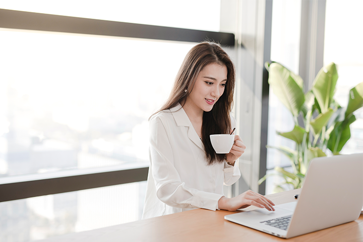 A young woman working on a laptop, smiling and holding a coffee mug.