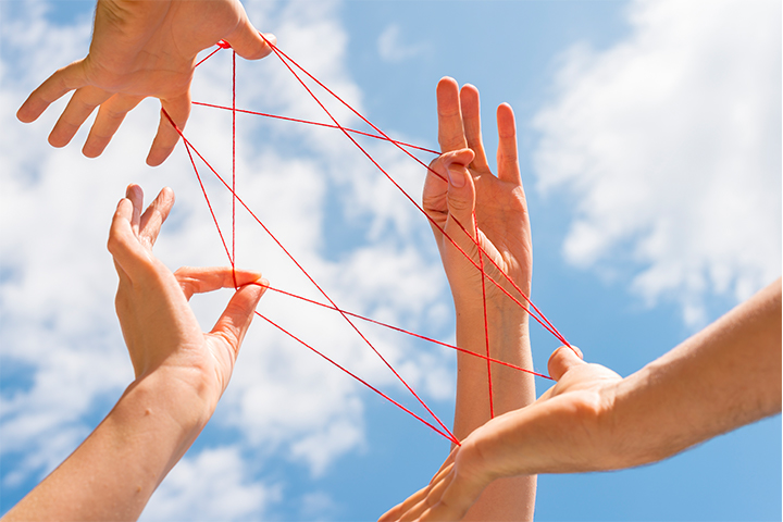 Two sets of hands working together with string to create a 'cat's cradle' design, under a blue sunny sky.