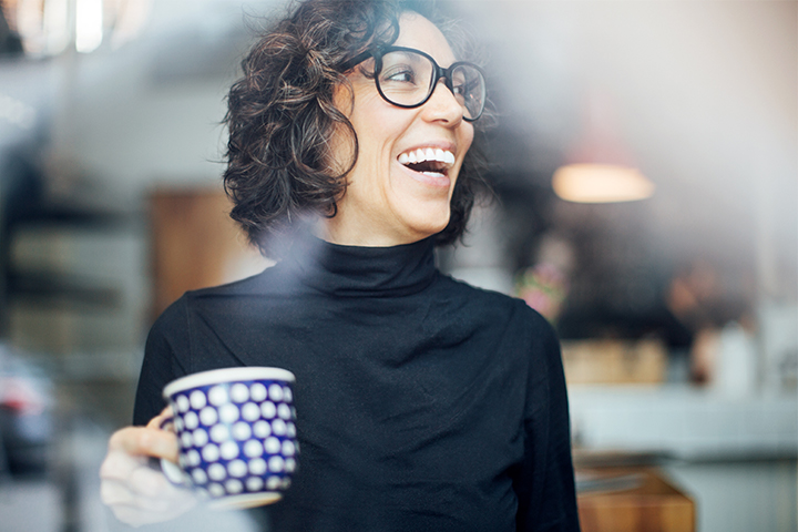 A young woman with curly hair and glasses, holding a coffee mug and smiling
