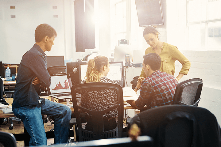 A group of 4 young professionals sitting around a computer collaborating, with sun streaming through the office windows.