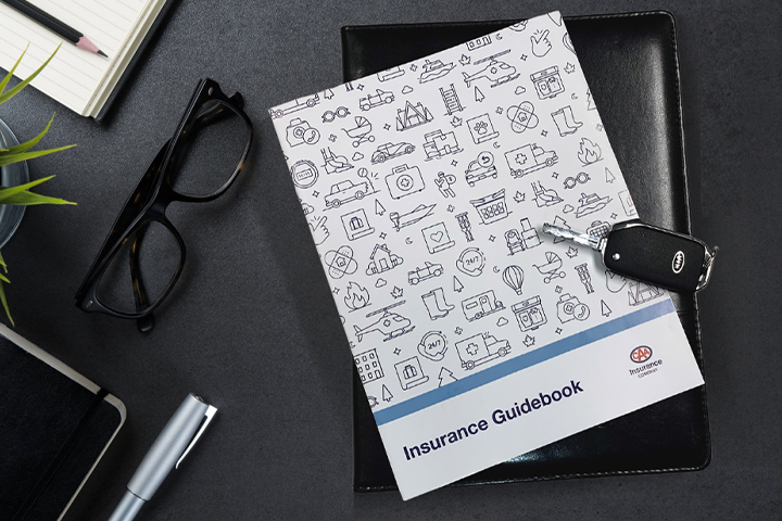 The CAA Insurance Guidebook lying on a desk next to a pair of glasses and a pencil and notepad, with a set of car keys lying on top.