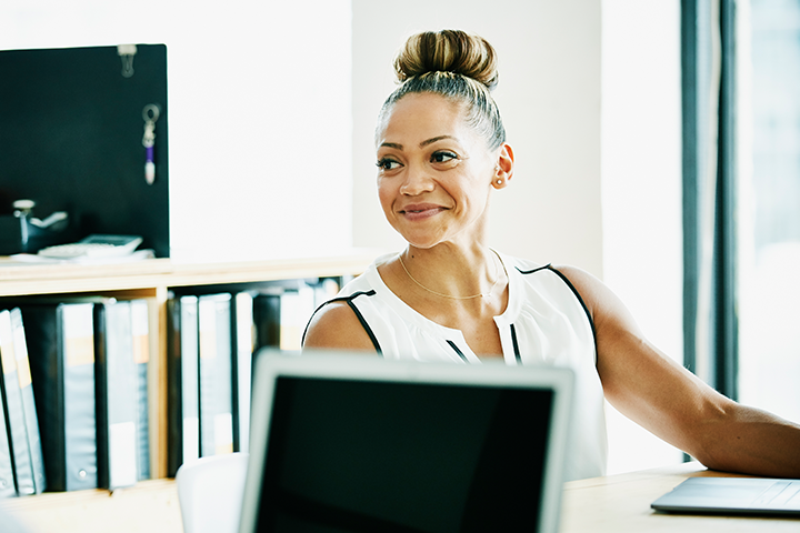 A professional woman sitting at a desk behind a laptop computer, looking to her right and smiling.