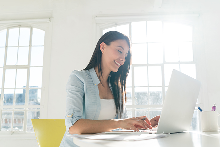 A young woman sitting at a desk typing a laptop computer, smiling.