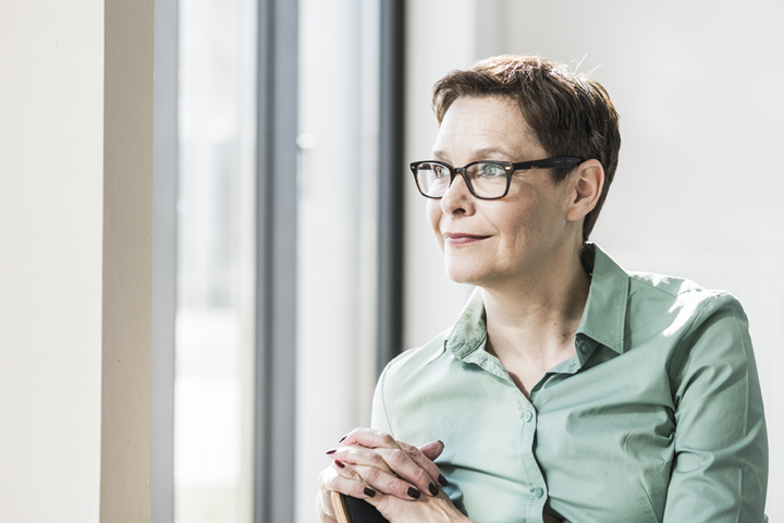 A mature business woman wearing a green shirt and glasses, sitting on a chair, looking out her office window.