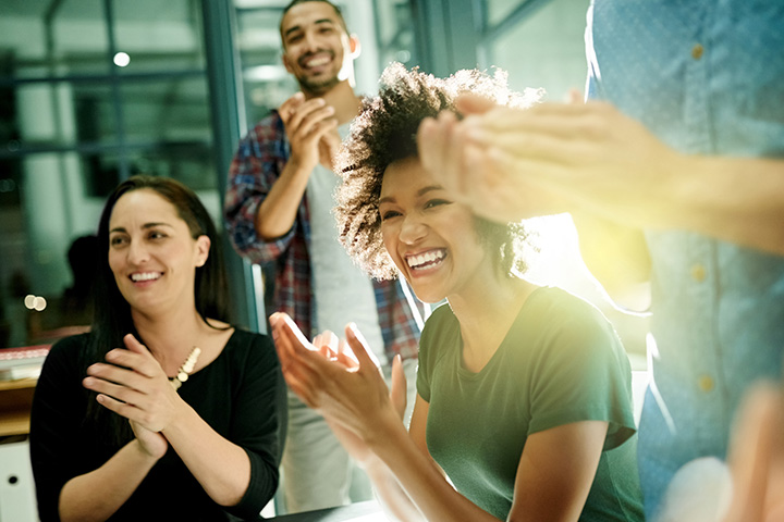 An office scene with people gathered around a table, all smiling and clapping.