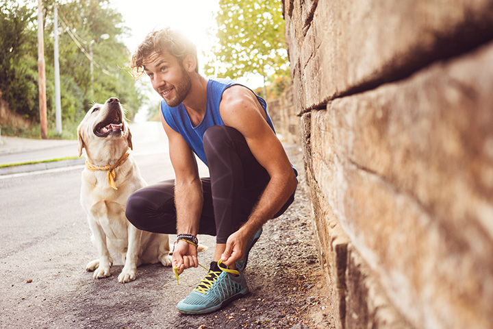 A young man kneeling down to tie his running shoe, with his dog (a blond lab) sitting next to him smiling.