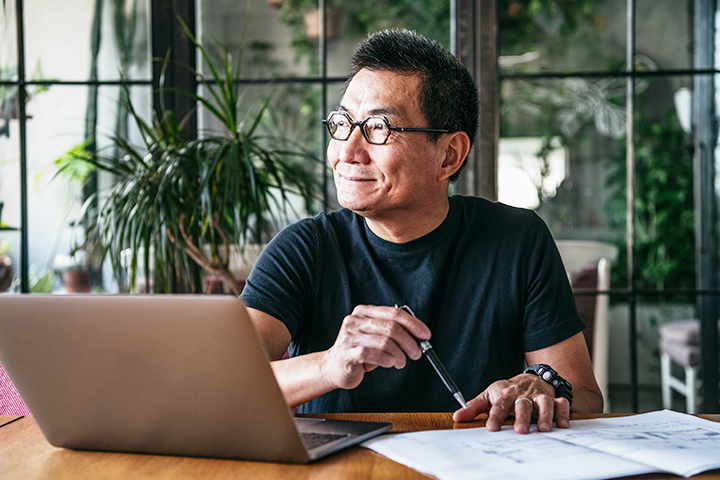 Middle aged man sitting at a desk in front of a lap top, looking out the window and smiling.