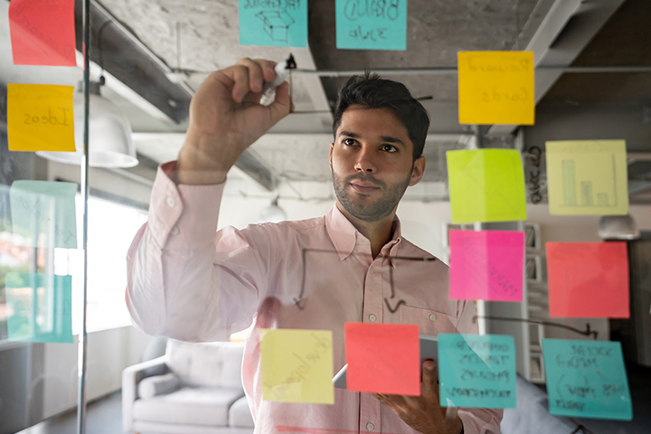 A man writing on a glass screen which is covered in coloured sticky notes.