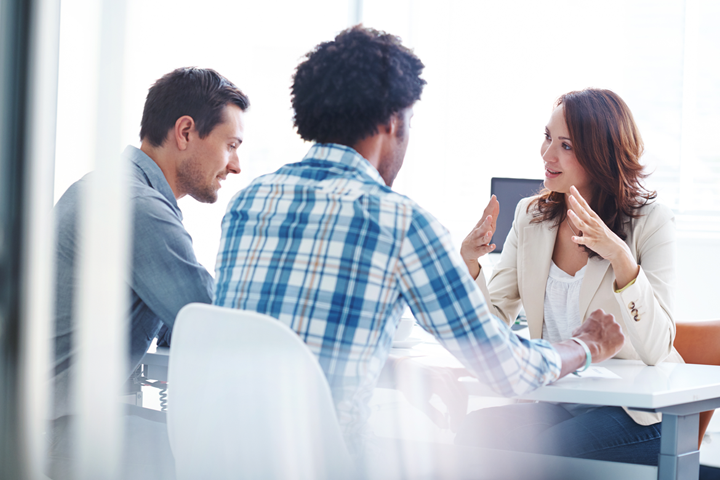 Two men sitting across a table from a professional woman, who is talking and using her hands to illustrate her point.