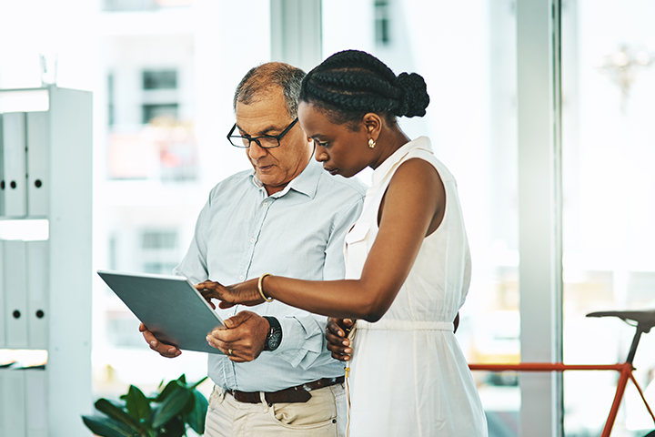 A man and a woman standing in an office, looking at a tablet with the woman pointing to the screen.