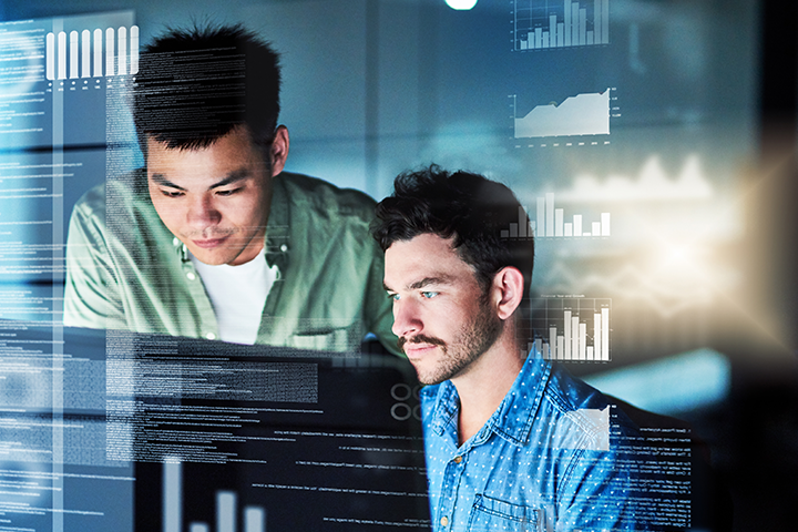 Young man sitting at a desk with another young man standing to his right, looking over his shoulder. Both are looking at a computer screen. Overlaying the image are various lines, graphs and charts depicting data.