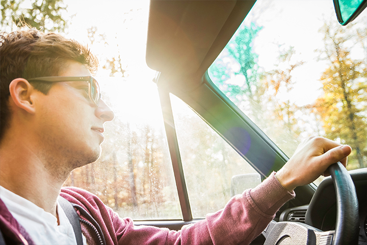 A young man driving a convertible on a country road surrounded by forest, with the sun shining through the trees.