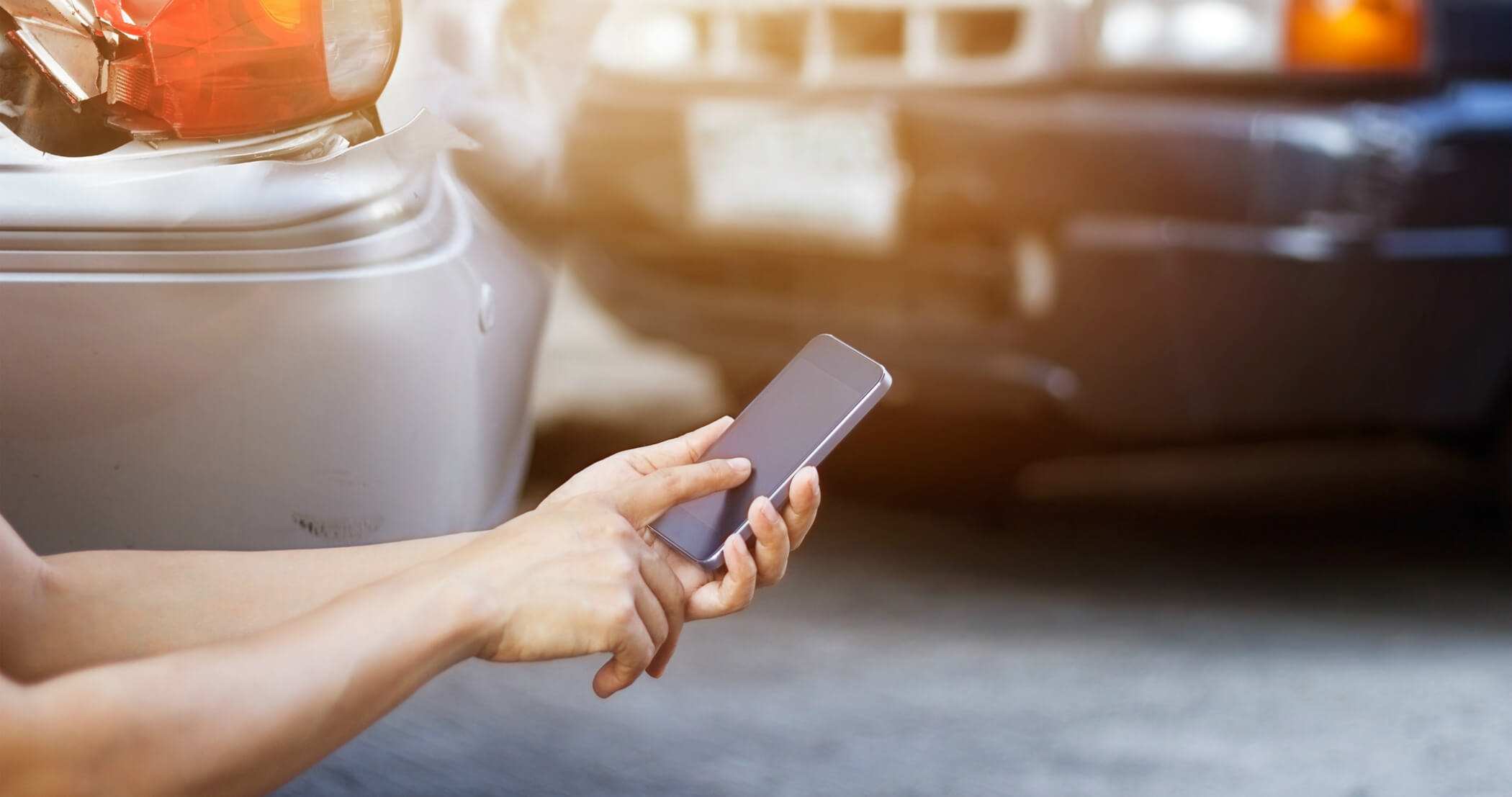 Woman holding a smartphone and dialing, in front of two vehicles that have been involved in an accident.