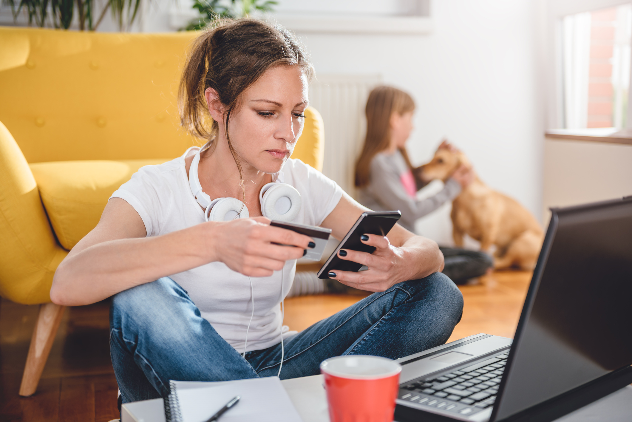 Woman sitting on the floor in front of a laptop, holding and looking at a smart phone in her left hand, and holding a credit card in her right hand. Young girl petting a golden retriever in the background.