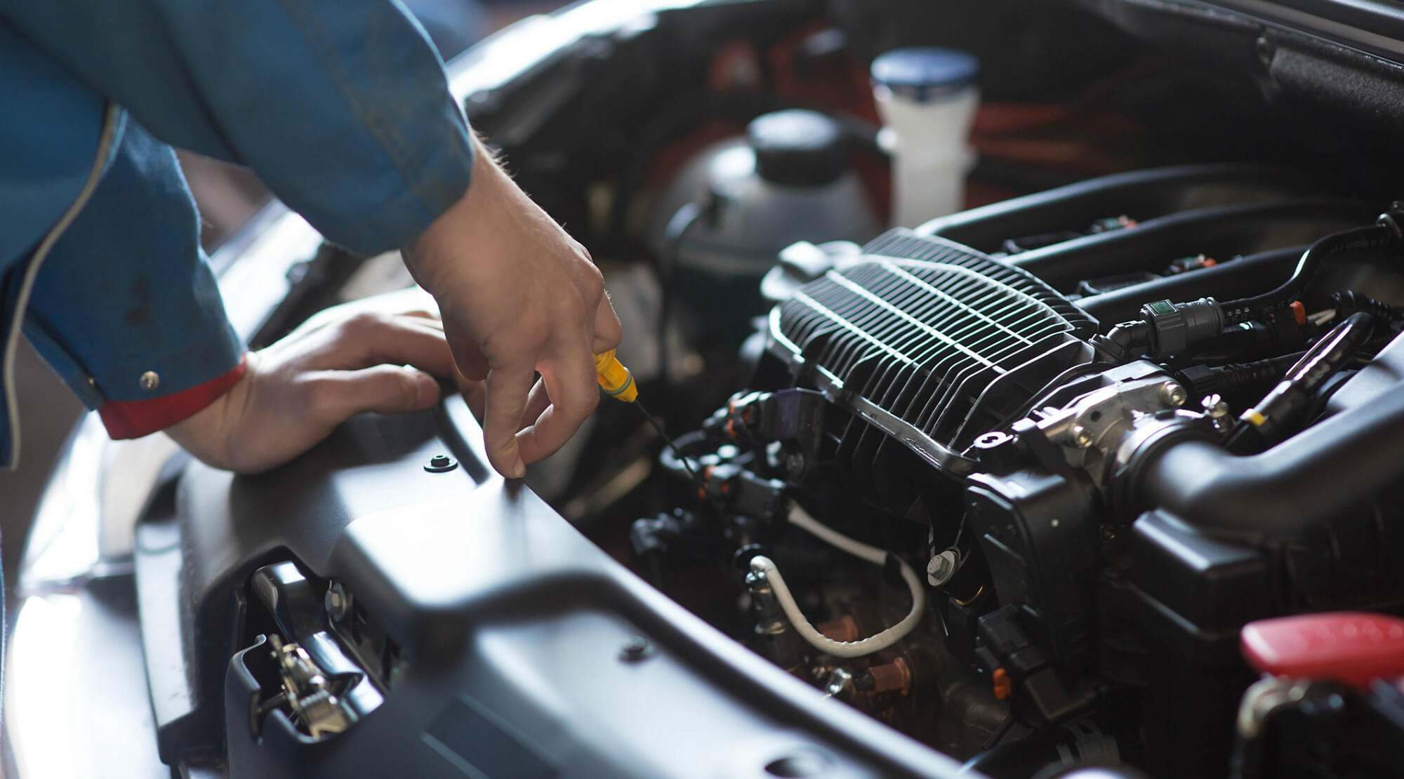 An open hood of a vehicle showing engine and other components, with a mechanic's hands holding a tool